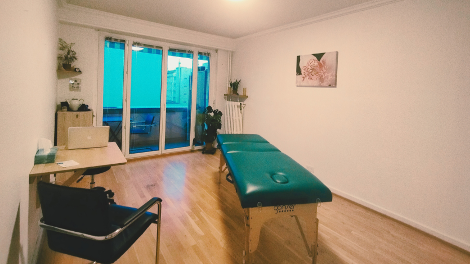 Salle de traitement L'espace thérapeutique est spacieux et épurée. Il y a un petit bureau et la table de soin prend l'espace pricinpal. Quelques plantes vertes et un tableau. Une grande baie vitrée prend tout le mur et laisse voir un blacon sans vis à vis.